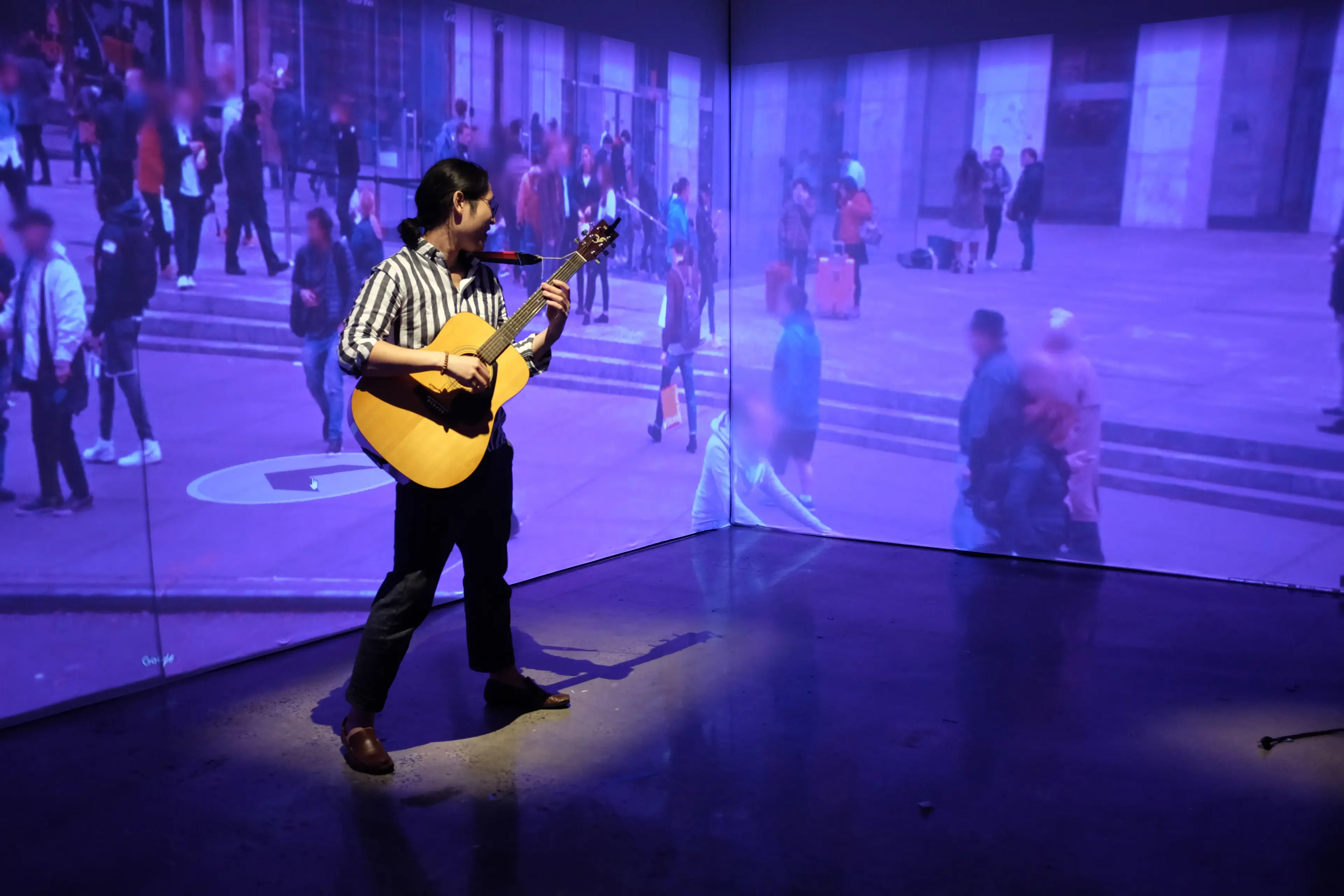 A guitarist performing inside the projection room, silhouetted against a wall-scale street scene of pedestrians, the projected environment wrapping around the floor and walls