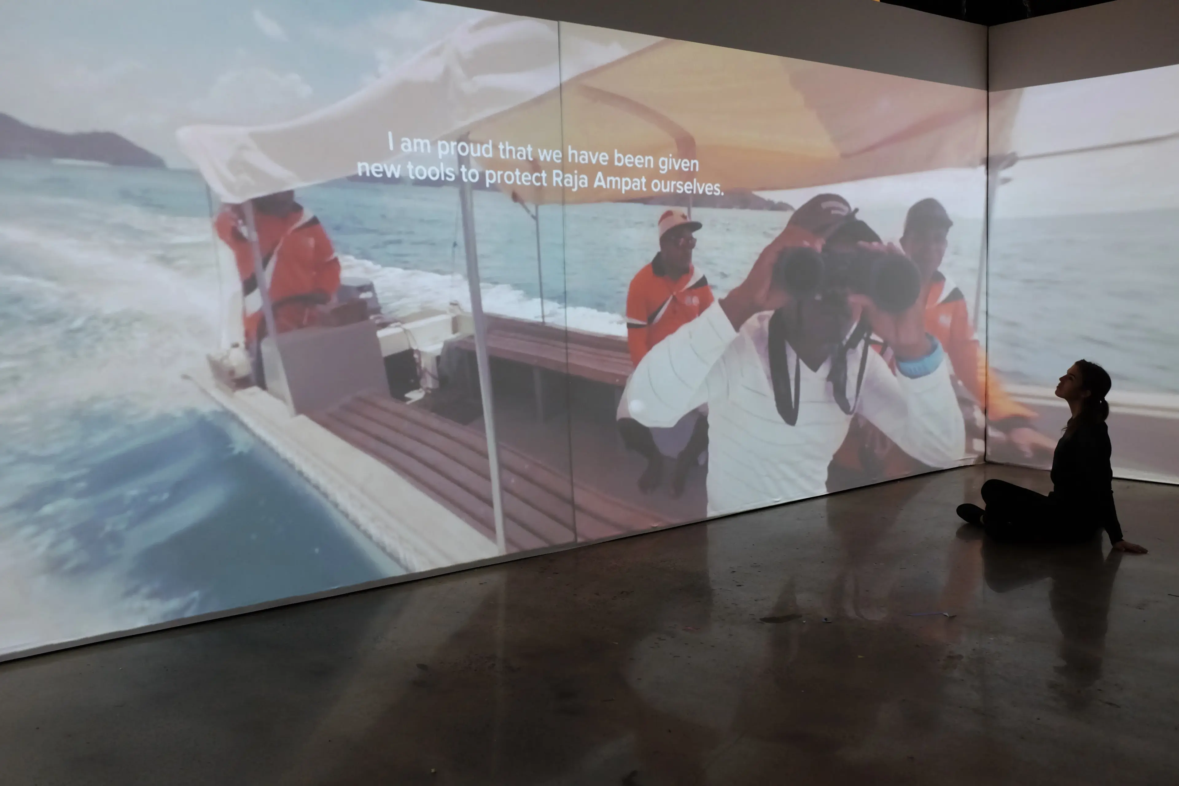 A person sitting on the floor of the projection room, looking up at a wall-spanning documentary image of people on a boat at sea with subtitled text overlay